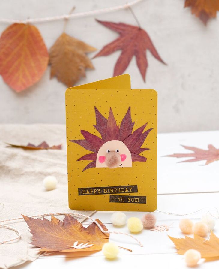 Auf einem hellen Untergrund, vor einer hellgrauen Betonwand, steht eine selbstgebastelte Igel-Klappkarte aus Herbstblättern und Materialien von Artoz. Drumherum und im Hintergrund sieht man viele getrocknete und gepresste Herbstblätter sowie kleine Mini-Pompons. Foto: Johanna Rundel