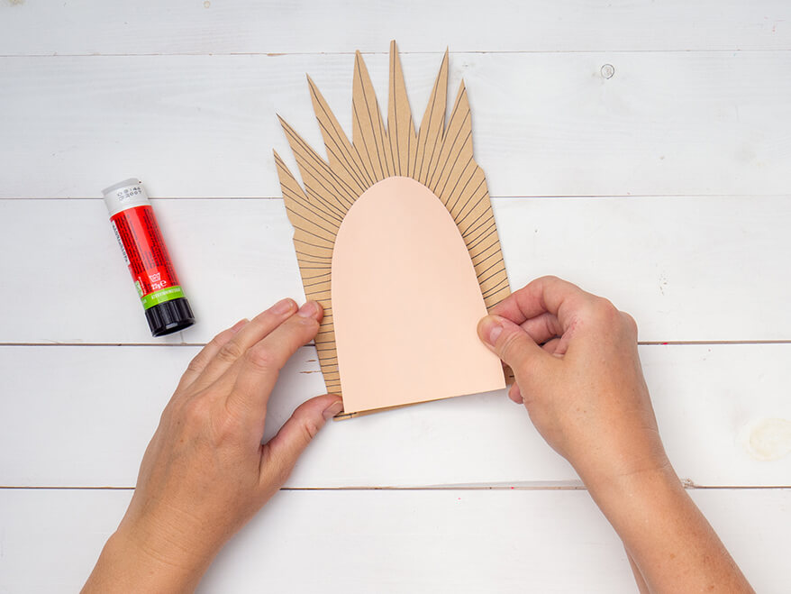 Jemand klebt einen länglichen Halbkreis aus rosa Tonkarton von Artoz auf eine braune Papiertüte mit Streifen und Zacken am oberen Rand. Der Untergrund ist weiß. Foto: Johanna Rundel