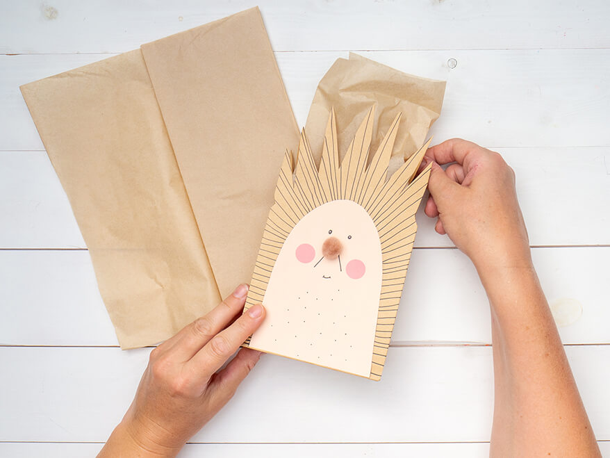 Jemand schlägt eine, zu einem Igel umgestaltete, braune Papiertüte mit einem hellbraunen Seidenpapier von Artoz aus. Der Untergrund ist weiß. Foto: Johanna Rundel
