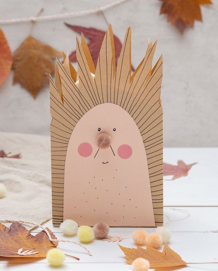 Auf einem hellen Untergrund, vor einer hellgrauen Betonwand, steht eine zu einem Igel umgestaltete, braune Papiertüte von Artoz. Drumherum und im Hintergrund sieht man viele getrocknete und gepresste Herbstblätter sowie Mini-Pompons in Herbstfarben. Foto: Johanna Rundel