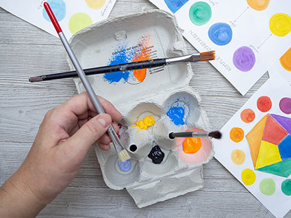 Auf einem hellgrauen Holzuntergrund steht ein leerer Eierkarton. In die Mulden wurden verschiedene Farben gegeben. Die Spitzen halten Pinsel. Eine Hand mit Pinsel ragt von links unten ins Bild und nimmt Farbe auf. Rechts neben dem Eierkarton liegen verschiedene gemalte Bilder.