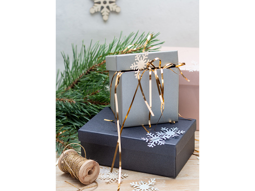 Auf einem Holztisch stehen drei Geschenkschachteln von Artoz. Eine schwarze, eine rosafarbene und eine graue. Der Deckel der grauen Schachtel ist leicht geöffnet und es schauen goldene Flitterfäden heraus. Auf den Schachteln sind Schneeflocken aus Papier verteilt. Neben und hinter den Schachteln ist eine Garnrolle sowie ein Tannenzweig zu sehen. Foto: Johanna Rundel