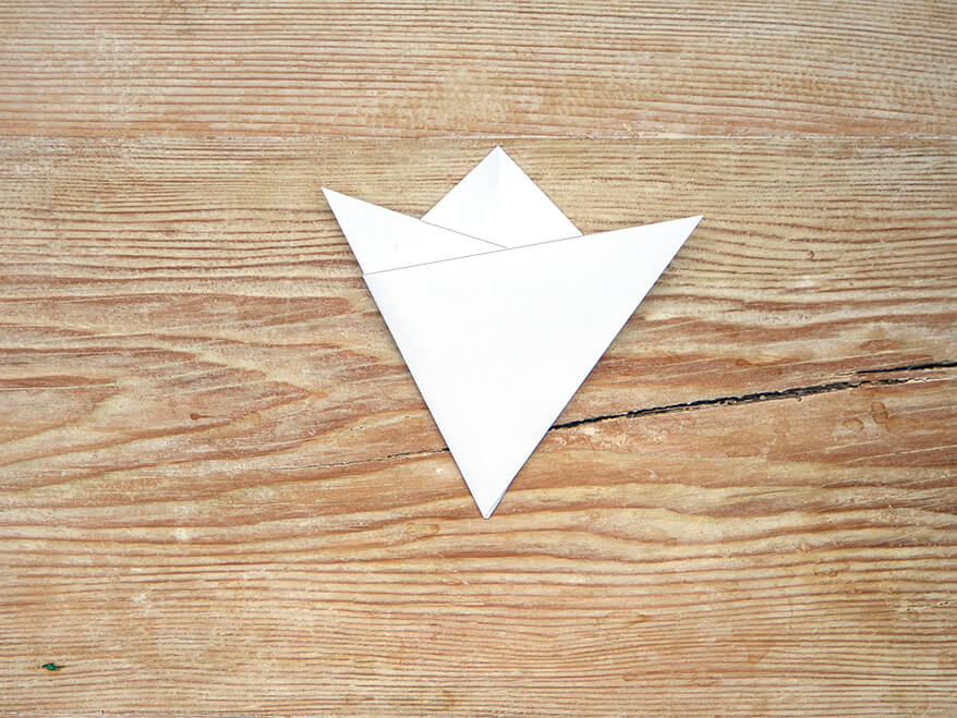 Ein, zu einem Dreieck gefaltetes quadratisches Blatt Papier der Marke Artoz. Beide Spitzen des Dreiecks wurde schräg nach oben gefaltet. Der Untergrund ist ein rustikales, helles Holzbrett. Foto: Johanna Rundel