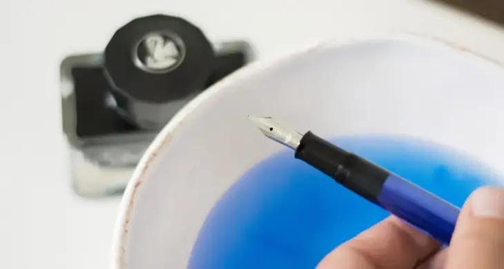 Ein offener Pelikan Füller über einer Schüssel mit Wasser, welches von der Tinte blau gefärbt ist. Neben der Schüssel steht ein Tintenfass der Marke Pelikan.