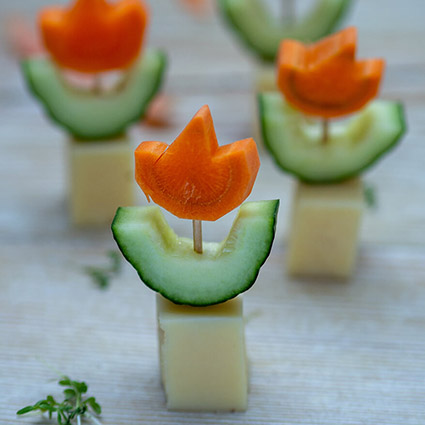 Ein Käsewürfel als Blumentopf, eine zu zwei Blättern zurechtgeschnittene Gurkenscheibe und eine Blüte aus Karotte wurden mit einem Zahnstocher zu einer Blume zusammengesteckt. Davon sieht man vier auf einem Holzbrett stehen. Foto: Johanna Rundel