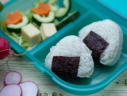 In einer türkisfarbenen Brotdose von School-Mood liegen im rechten Fach zwei Reispäckchen (Onigiri) und im linken Käse-Rohkostspieße in Blumenform. Vor der Dose liegen ein paar Radieschenscheiben. Alles liegt auf einem Holzbrett. Foto: Johanna Rundel
