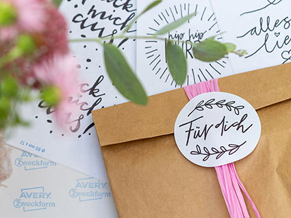 Auf einem Tisch liegen mehrere runde Blanko-Etikettbögen von Avery Zweckform verteilt, die mit unterschiedlichen Geschenkaufkleber-Designs bedruckt wurden. Auf den Druckbögen liegt eine braune Papiertüte um die ein rosa Band gewickelt wurde. Darauf wurde ein runder Aufkleber mit der Aufschrift „Für dich“ geklebt. Im Vordergrund ragt unscharf ein Blumenstrauß ins Bild. Foto: Johanna Rundel