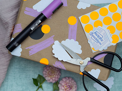 Auf einem türkisfarbenen Untergrund liegt ein in Kraftpapier eingepacktes Geschenk. Das Papier wurde mit verschiedenen Aufklebern von Avery Zweckform verschönert und mit einem lila Stift bemalt. Auf dem Geschenk liegen weitere Aufkleberbögen mit Markierungspunkten, eine schwarze Schere und ein lila Stift. Eine rosafarbene Tischdecke und zwei Dahlienblüte sind an den Rändern des Bildes zu sehen. Foto: Johanna Rundel