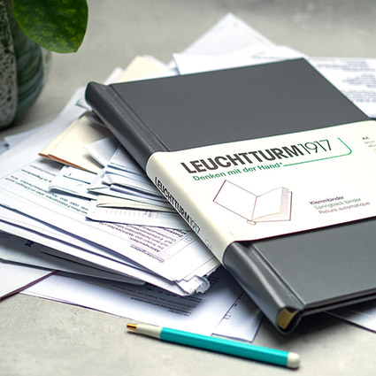 Auf einem hellgrauen Untergrund liegt ein Stapel aus Unterlagen. Auf den Unterlagen ist ein dunkelgrauer Klemmbinder von Leuchtturm1917 zu sehen. Vor dem Stapel liegt ein türkisfarbener Kugelschreiber und dahinter steht eine Topfpflanze. Foto: Johanna Rundel