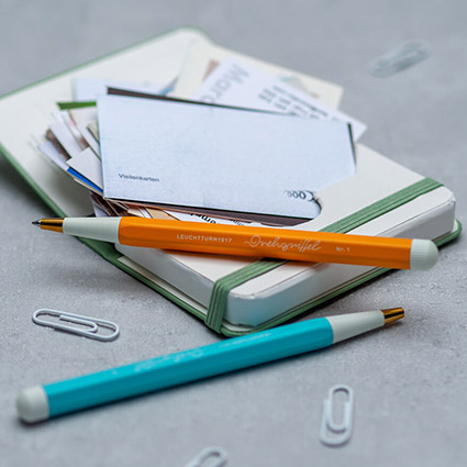 Auf einem hellgrauen Untergrund liegt eine salbeifarbenes Visitenkartenetui von Leuchtturm1917, das mit Visitenkarten gefüllt ist. Darauf und daneben liegen ein orangefarbener und ein türkiser Drehgriffel. Einige weiße Büroklammern liegen verstreut darum. Foto: Johanna Rundel