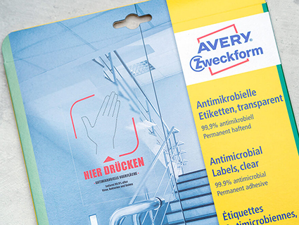 Auf einem grauen Untergrund in Betonoptik liegt eine Packung transparente, antimikrobielle Etiketten von Avery-Zweckform. Auf der Verpackung ist ein Treppenhaus mit Glastür abgebildet, auf der Glastür klebt ein Aufkleber auf dem „Bitte hier drücken – antimikrobielle Oberfläche“ steht. Foto: Johanna Rundel
