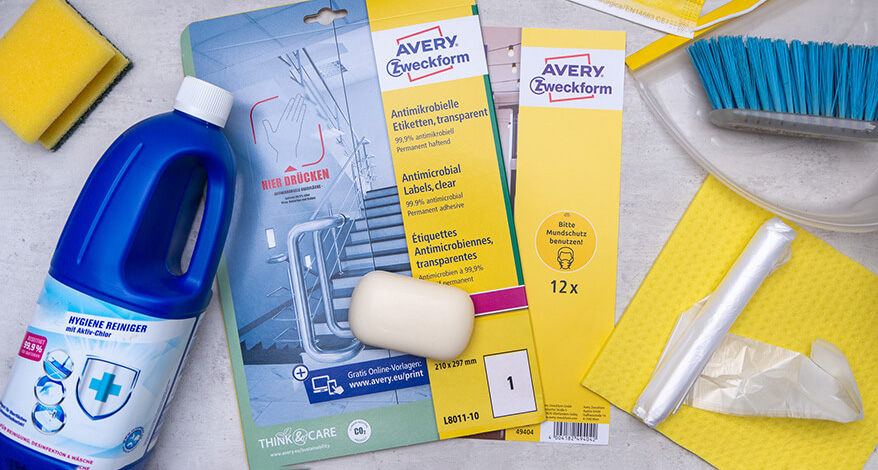Auf einem grauen Untergrund in Betonoptik liegen eine Packung Hinweis-Aufkleber zum Thema Mundschutz und eine Packung antimikrobielle Etiketten von Avery-Zweckform. Drumherum sind verschiedene Putzutensilien zu sehen: eine blaue Flasche Hygienereiniger, ein gelber Spülschwamm und ein gelbes Schwammtuch, ein Kehrblech und -besen, ein weißes Seifenstück, Müllbeutel sowie gelbe Mund-Nase-Bedeckungen. Foto: Johanna Rundel
