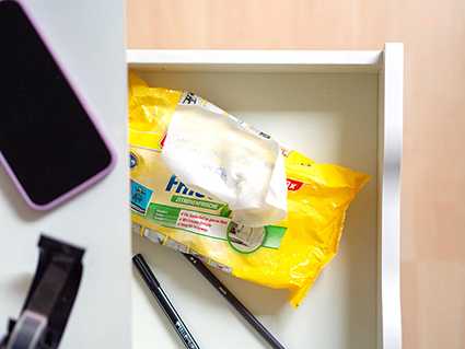 In einer ausgezogenen, weißen Schreibtischschublade liegt eine geöffnete, gelbe Packung mit Mehrzweckreinigungstüchern. Daneben liegen zwei Stifte. Auf dem weißen Schreibtisch ist ein Smartphone und eine Klebebandabroller zu sehen. Foto: Johanna Rundel