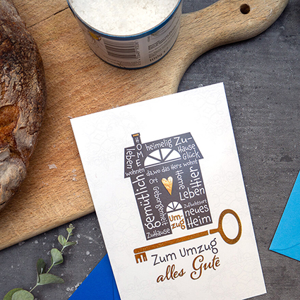 Auf einem anthrazitfarbenen Untergrund liegt eine Grußkarte des ABC Glückwunschkartenverlags, darunter ein blauer Briefumschlag. Auf der Karte sind ein schwarzes Haus und ein goldener Schlüssel abgebildet. Unter dem Schlüssel steht in goldenen Buchstaben: „Zum Umzug alles Gute“. Über der Karte ist ein Holzbrett zu sehen, auf dem Brot und Salz stehen. Foto: Johanna Rundel