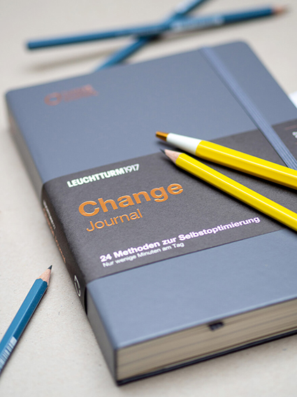 Auf einem grauen Untergrund liegt das blaue Change Journal von Leuchtturm1917. Auf dem Journal liegen ein gelber Drehgriffel und ein gelber Bleistift. Weitere blaue Bleistifte sind um das Journal verteilt. Foto: Johanna Rundel