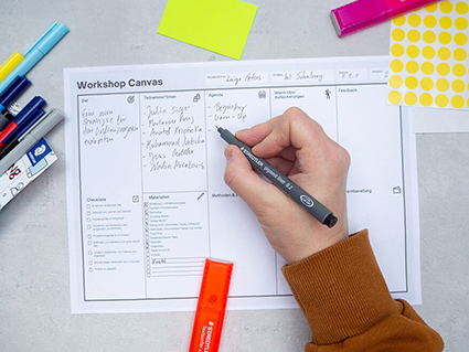 Jemand schreibt mit einem schwarzen Stift von Staedtler auf einen Workshops-Canvas. Drumherum liegen verschiedene Workshopmaterialien, wie Textmarker, Haftnotizzettel und Klebepunkte. Der Untergrund ist hellgrau. Foto: Johanna Rundel