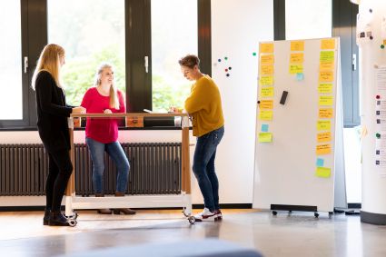 Drei Frauen stehen an einem Stehtisch und reden miteinander. Rechts neben ihnen steht eine mobiles Whiteboard, das mit zahlreichen bunten Haftnotizen beklebt ist, welche beschriftet sind. Im Hintergrund ist verschwommen ein Fenster zu sehen.