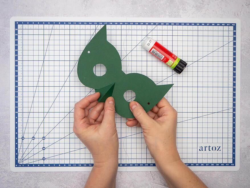 Jemand klebt einen Schnabel in den Ausschnitt einer dunkelgrünen Vogelmaske aus Fotokarton. Der Untergrund ist eine helle, faltbare Schneidematte mit Linien von Artoz. Foto: Johanna Rundel