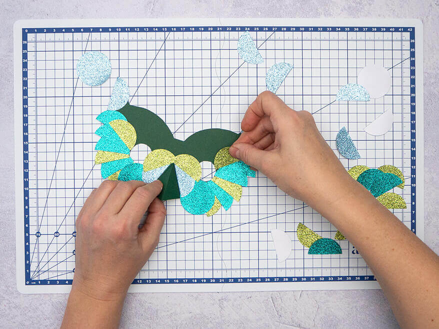 Jemand klebt Halbkreise aus verschiedenen Papieren in Grün- und Blautönen auf eine dunkelgrüne Vogelmaske aus Papier. Der Untergrund ist eine helle, faltbare Schneidematte mit Linien von Artoz. Foto: Johanna Rundel