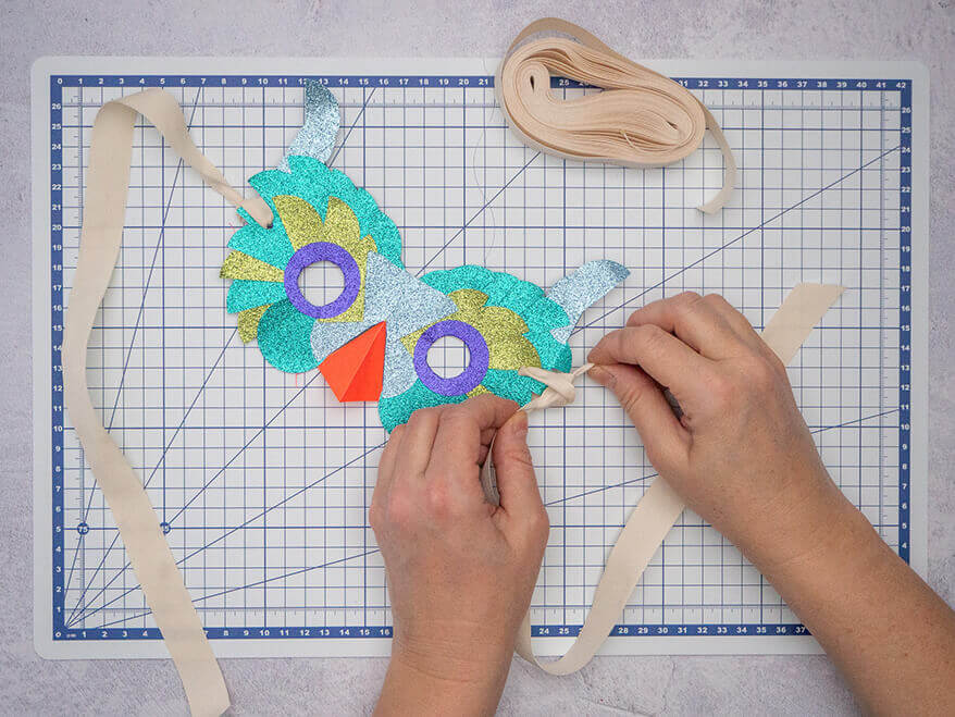 Jemand knotet Bänder an die seitlichen Löcher in einer bunten Vogelmaske aus Papier in verschiedenen Grün- und Blautönen. Der Untergrund ist eine helle, faltbare Schneidematte mit Linien von Artoz. Foto: Johanna Rundel