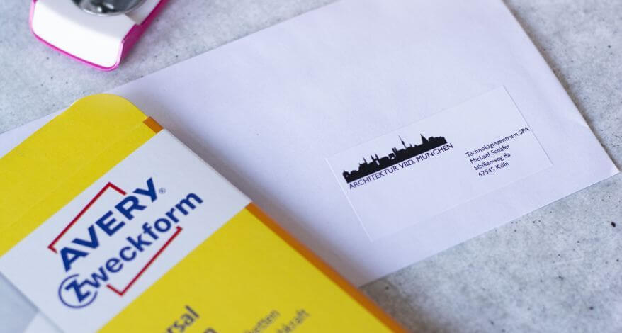 Nahaufnahme eines Briefumschlags mit einem Adressetikett. Auf dem Etikett ist die Stadtsilhouette von München aufgedruckt, darunter steht „Architektur Vbd. München“. Rechts unten steht auf dem Etikett der Empfänger. Links ragt die Verpackung der verwendeten Adressetiketten von Avery Zweckform ins Bild.