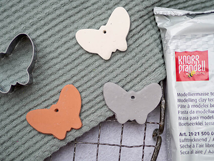 Auf einem Kuchengitter liegt ein salbeigrünes Küchenhandtuch. Darauf liegen drei, aus Modelliermasse ausgestochene, Schmetterlinge in Terracotta, Weiß und Betonoptik. Rechts daneben liegt eine Packung lufttrocknende Modelliermasse von Knorr Prandell. Foto: Johanna Rundel