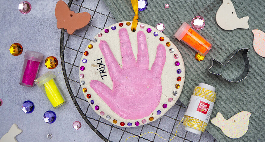 Auf einem Kuchengitter liegt ein salbeigrünes Küchenhandtuch. Darauf liegt ein Handabdruck in einem Salzteig-Kreis, der mit bunten Glitzersteinen von Knorr Prandell verziert wurde. Neben der Hand steht in schwarzer Schrift "TRIXI". Um das Salzteigobjekt herum liegen verschiedene Bastelmaterialien, wie Glitzersteine, Glitterdöschen, Keksausstecher und bereits ausgestochene Vögel aus Modelliermasse. Der Untergrund ist Betongrau. Foto: Johanna Rundel