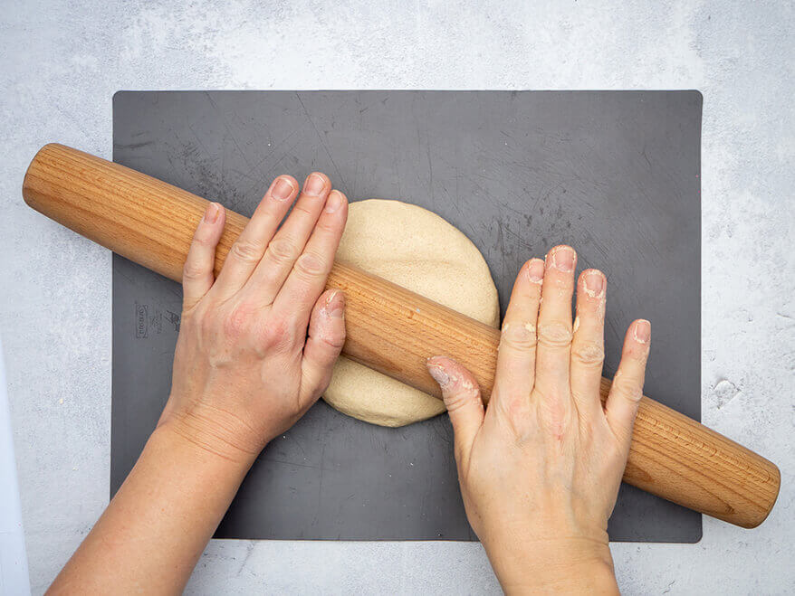 Jemand rollt auf einer dunkelgrauen Backmatte mit einem Nudelholz Salzteig zu einem Kreis aus. Der Untergrund ist Betongrau. Foto: Johanna Rundel