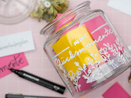 Auf einem rosafarbenen Untergrund mit weißem Karomuster liegt ein Bonbonglas, auf das mit einem weißen Paintmarker von Staedtler „Meine Glücksmomente“ geschrieben wurde. Unter dem Schriftzug sind florale Ornamente gezeichnet. Im Glas befinden sich gelbe und pinke Zettel. Einige Zettel liegen lose um das Glas verteilt. Außerdem ist der Deckel des Glases, sowie Stifte und ein paar Hortensienblüten im Bild zu sehen. Foto: Johanna Rundel