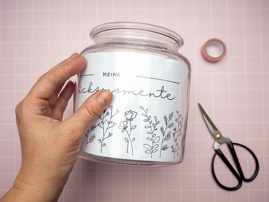 Jemand klebt mit Washitape einen Vorlagebogen zum Abpausen in das Innere eines Bonbonglases. Der Untergrund ist ein rosafarben mit weißem Karomuster. Foto: Johanna Rundel