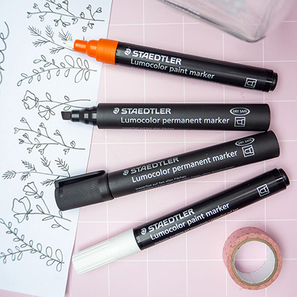 Auf einem rosafarbenen Untergrund mit weißem Karomuster liegen aufgefächert vier schwarze Stifte von Staedtler: zwei Permanentmarker und zwei Paintmarker. Unter den Stiften liegt ein Blatt mit einem floralen Muster zum Abpausen. Daneben ist eine Rolle Washitape zu sehen. Foto: Johanna Rundel