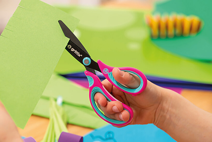 Eine Kinderhand schneidet mit einer pink-türkisfarbenen Griffix-Schere von Pelikan ein grünes Papier zurecht. Im Hintergrund sieht man unscharf weitere Papiere auf einem Tisch liegen.