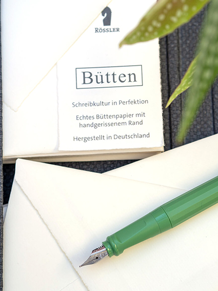 Es ist ein Ausschnitt einer schwarzen Mappe zu sehen, in der Bütten-Briefpapier und Butten-Umschläge von Rössler stecken. Auf dem Stapel mit Umschlägen liegt ein hellgrüner Füller. In der rechten oberen Ecke ragen ein paar Blätter einer Zimmerpflanze ins Bild. Foto: Johanna Rundel