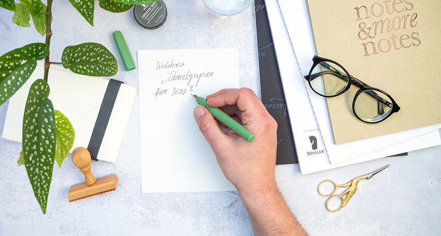 Hand schreibt mit grünem Füller auf Briefpapier, Schreibutensilien liegen daneben.