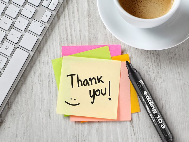 Auf einer hellen Holzoberfläche liegt ein Stapel bunter Haftnotizen. Auf dem obersten Zettel wurde mit einem schwarzen edding Marker "Thank You!" geschrieben und ein Smiley gemalt. Der Marker liegt daneben, ebenso wie die Tastatur eines Laptops. In der oberen rechten Ecke ist eine mit Kaffee gefüllte Tasse zu sehen.