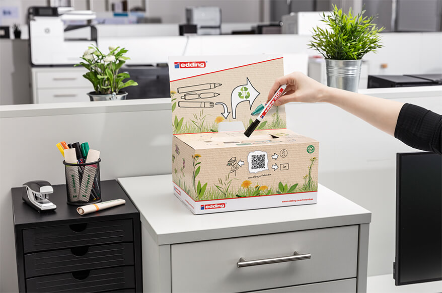 Jemand steckt einen Marker in die Recycling-Rückgabebox von edding, die auf einem niedrigen weißen Aktenschrank steht. Im Hintergrund sieht man ein Großraumbüro.