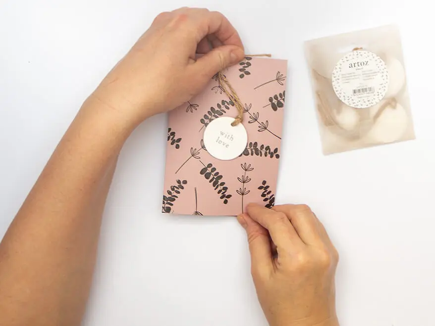 Jemand befestigt einen runden, weißen Geschenkanhänger mit der Aufschrift "with Love" von Artoz an einer mit botanischen Motiven bestempelten, altrosafarbenen Klappkarte. Der Untergrund ist weiß. Foto: Johanna Rundel