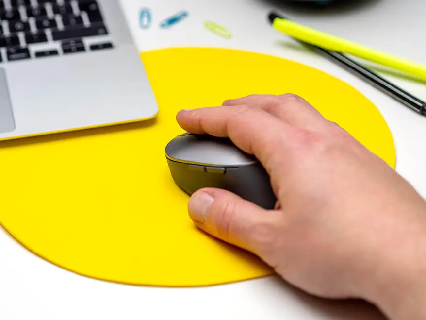 Jemand bedient eine schwarz-graue Computermaus mit der rechten Hand. Die Maus liegt auf einem gelben Mousepad, das aus Moosgummi von Knorr Prandell selbst gebastelt wurde. Im Hintergrund sieht man den Laptop, sowie ein paar Stifte und Büroklammern auf dem Schreibtisch. Foto: Johanna Rundel