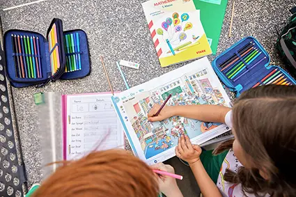Zwei Kinder im Grundschulalter machen auf einer Tischtennisplatte Schulaufgaben. Ein Lehrbuch ist aufgeschlagen. Das Mädchen schreibt etwas hinein. Die Mäppchen von ergobag liegen offen herum.