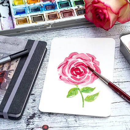 Eine weiß-pinke Rose liegt auf einer Aquarellpostkarte, auf der ebenfalls eine Rose aquarelliert wurde. Die Postkarte liegt auf einem schwarzen Sketchbook von Hahnemühle.