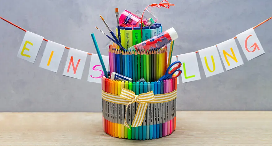 Eine Torte aus verschiedenen Stiften, Schulmaterialien und Markern der Marke Stadtler steht auf einem Tisch. Dahinter ist eine weiße Buchstaben-Girlande mit dem Wort „Einschulung“ aufgehängt.