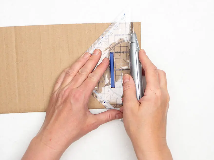 Jemand schneidet mit einem Cutter und einem Geodreieck von Staedtler ein Stück Pappe in Streifen.