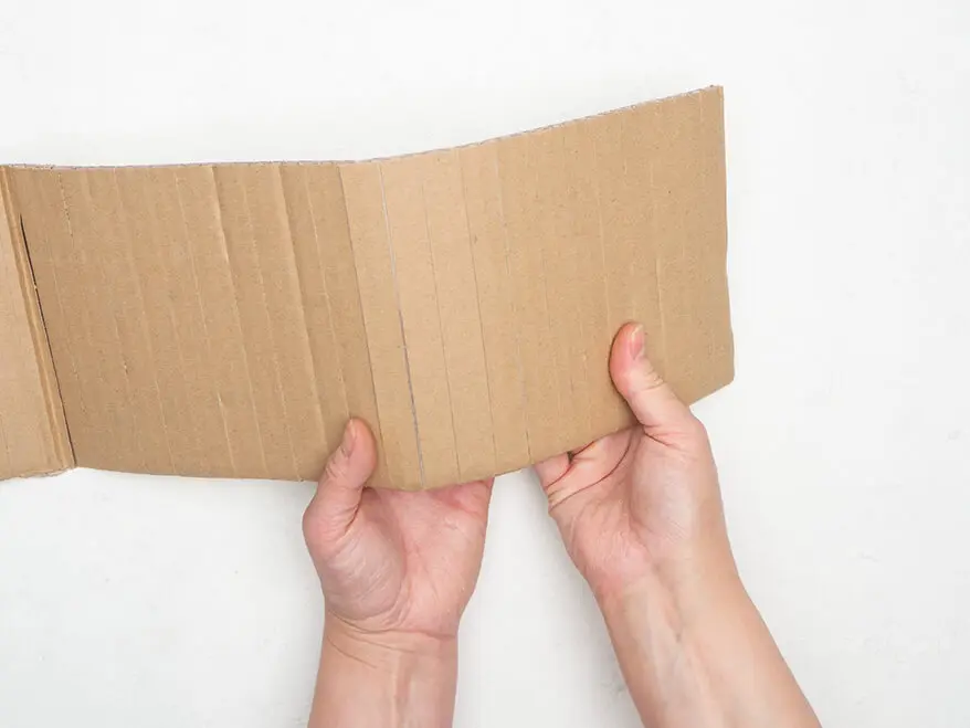 Jemand biegt einen zuvor vielmals senkrecht eingeritzten Streifen Pappe in eine Rundung.