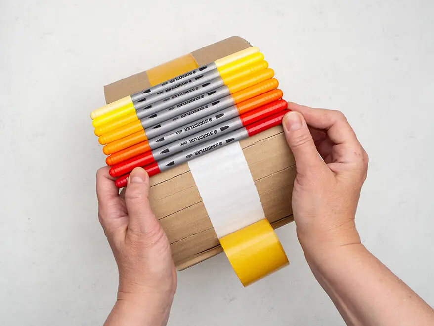 Jemand klebt viele bunte Fasermaler von Staedtler auf einen Streifen doppelseitiges Klebeband am Außenrand eines Pappkreises.