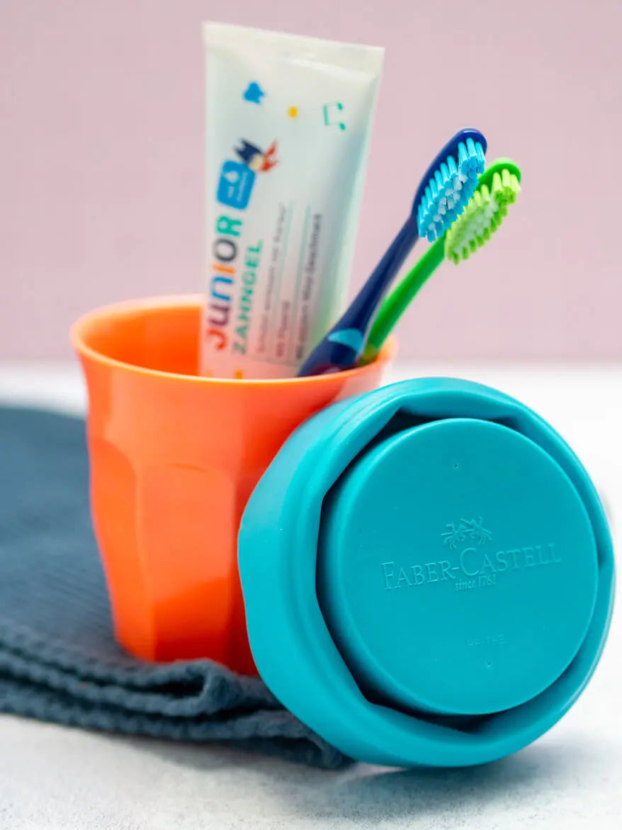 Ein orangener Zahnputzbecher mit Zahnbürsten und Zahnpasta steht auf einem blauen Handtuch. Foto: Johanna Rundel