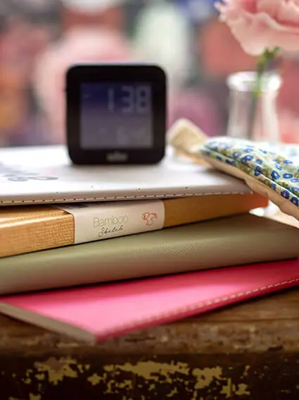 Verschiedener Skizzen- und Notizbücher von Hahnemühle stapeln sich auf einem rustikalen Hocker. Obendrauf steht ein Wecker, daneben liegt eine Schlafmaske.