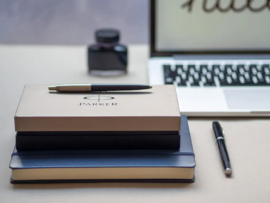 Auf einem hellgrauen Schreibtisch liegt ein blaues Notizbuch, auf dem ein Geschenketui von Parker steht. Foto: Johanna Rundel