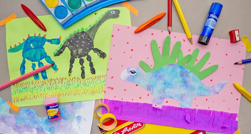 Auf einem grauen Untergrund liegen zwei, aus Handabdrücken selbstgestaltete, Dinosaurier-Bilder. Drumherum sind verschiedene Bastelmaterialien und Farbstifte von Pelikan und Herlitz verteilt. Foto: Johanna Rundel