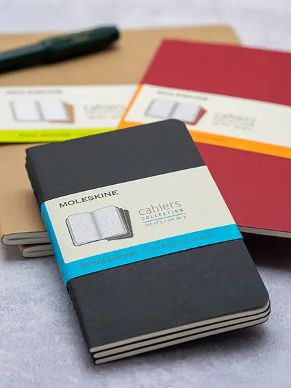 Drei Dreierpacks Notizhefte von Moleskine in Schwarz, Rot und Hellbraun liegen auf einem grauen Untergrund. Foto: Johanna Rundel