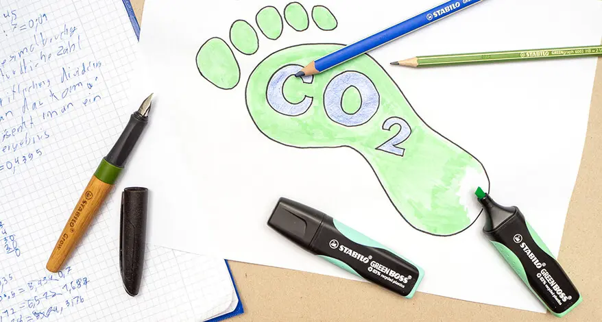 Auf einem grauen Untergrund liegt ein Rechenheft und ein weißes Blatt Papier, auf das ein CO²-Fußabdruckgezeichnet wurde. Verschiedene, umweltfreundliche Stifte von STABILO sind darauf verteilt. Foto: Johanna Rundel